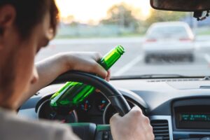 A young man holding a bottle of beer while driving, symbolizing the dangers of drinking and driving. DUI awareness and the risks of impaired driving.