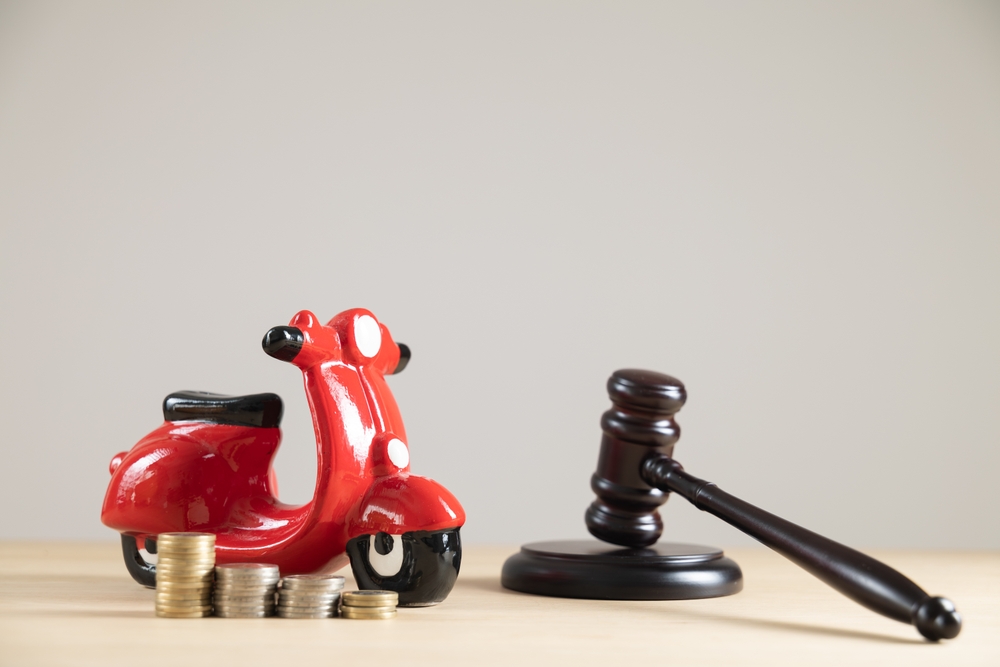 Close-up of a red motorbike-shaped piggy bank with a judge's gavel and cash on a plain background.