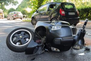 A motor scooter involved in a road accident, lying on its side near skid marks, with a damaged vehicle in the background. 