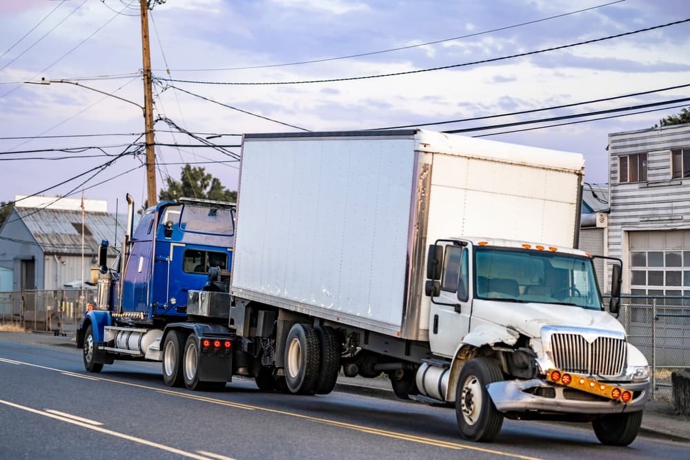 Mobile blue roadside assistance towing a powerful big rig semi truck tractor with a broken day cab semi truck and box trailer driving on a local road.