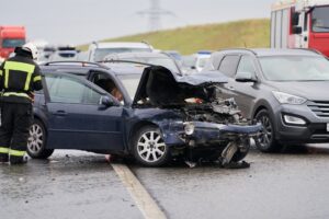 Consequences of a road accident. A car with a damaged hood and vegetables scattered on the engine. Nearby, a rescue worker tends to the injured driver.