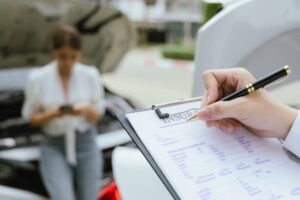 A young Caucasian man and an Asian woman inspect a damaged car following an auto accident. Nearby, an insurance agent assesses the vehicle and writes a detailed report.