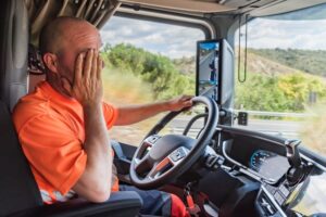 A truck driver sitting in the cab, rubbing his face with one hand, displaying visible fatigue and exhaustion as he drives the truck. 