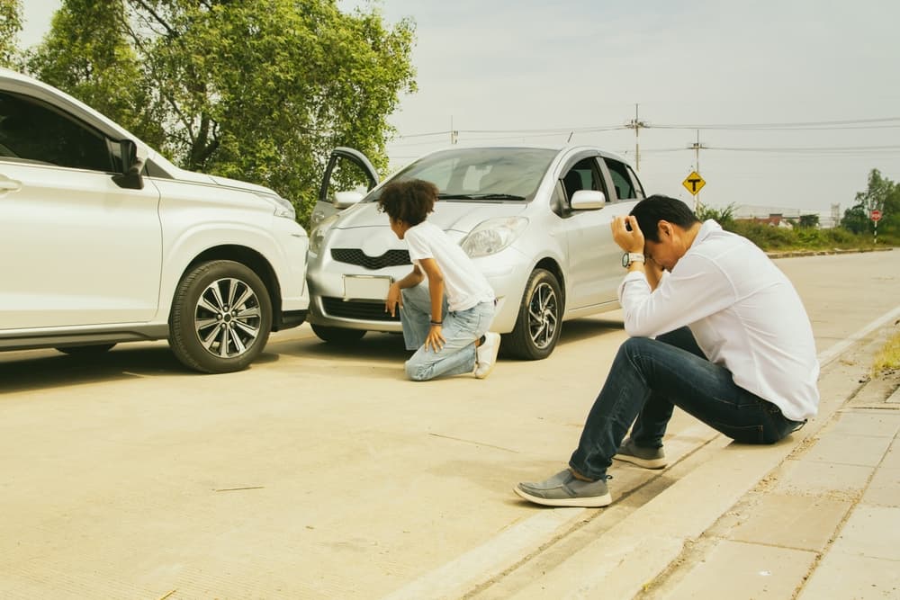 A man and a woman sit stressed after a serious car accident, with visible damage to the front bumper, as they wait for the insurance company. The crash resulted from illegal driving or traffic violations.