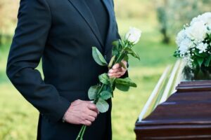 Close-up of an elderly widower in a black suit holding white roses, standing solemnly by his wife's coffin at her funeral.