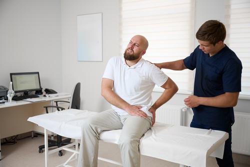 Man seated on exam table clutching hip while clinician supports shoulder