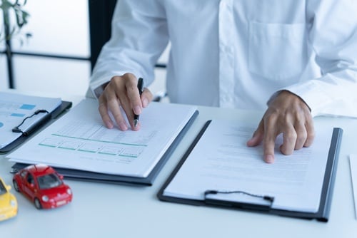 Person signing clipboard forms beside toy cars on desk during insurance paperwork