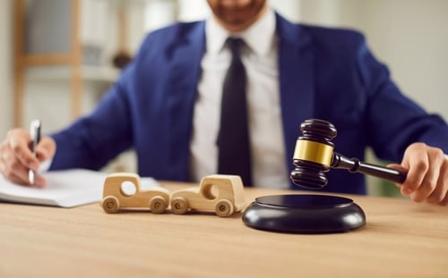 suited attorney holding gavel beside wooden toy cars on desk while writing