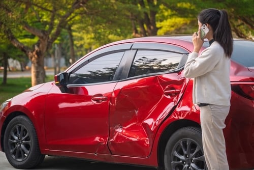 woman on phone standing beside car with visible collision damage